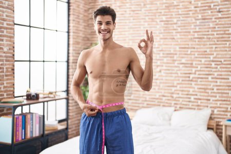 Foto de Young hispanic man using tape measure measuring waist doing ok sign with fingers, smiling friendly gesturing excellent symbol - Imagen libre de derechos