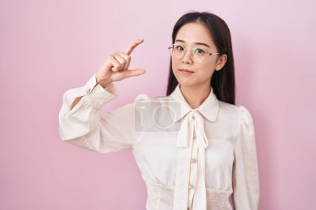Photo for Young chinese woman standing over pink background smiling and confident gesturing with hand doing small size sign with fingers looking and the camera. measure concept. - Royalty Free Image