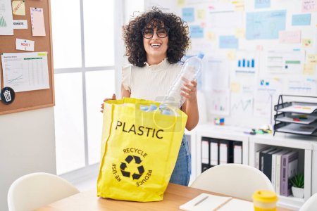 Téléchargez les photos : Jeune femme du Moyen-Orient tenant sac de recyclage avec des bouteilles en plastique au bureau souriant et riant fort parce que drôle blague folle. - en image libre de droit