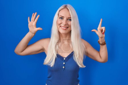 Foto de Mujer caucásica de pie sobre fondo azul mostrando y señalando hacia arriba con los dedos número siete mientras sonríe confiado y feliz. - Imagen libre de derechos