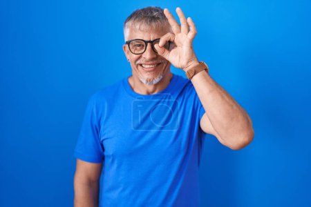 Photo for Hispanic man with grey hair standing over blue background doing ok gesture with hand smiling, eye looking through fingers with happy face. - Royalty Free Image