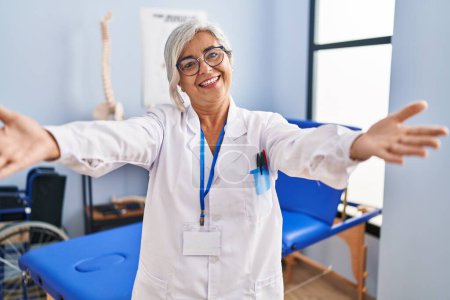 Téléchargez les photos : Middle age woman with grey hair working at pain recovery clinic looking at the camera smiling with open arms for hug. cheerful expression embracing happiness. - en image libre de droit