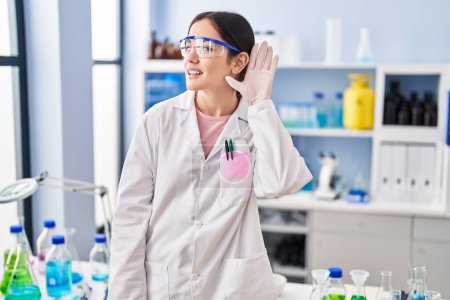 Photo for Young brunette woman working at scientist laboratory smiling with hand over ear listening and hearing to rumor or gossip. deafness concept. - Royalty Free Image