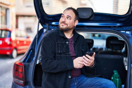Téléchargez les photos : Jeune homme hispanique utilisant smartphone assis sur le coffre de la voiture dans la rue - en image libre de droit