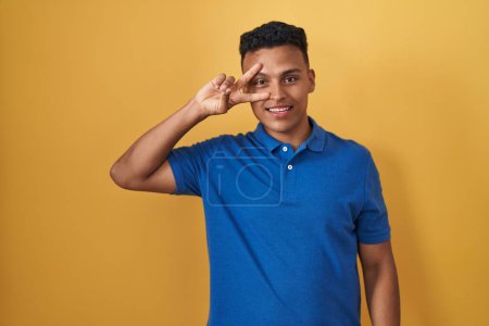 Foto de Joven hombre hispano de pie sobre fondo amarillo haciendo símbolo de paz con los dedos sobre la cara, sonriendo alegre mostrando la victoria - Imagen libre de derechos