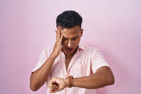 Foto de Joven hombre hispano de pie sobre fondo rosa mirando la hora del reloj preocupado, con miedo de llegar tarde - Imagen libre de derechos