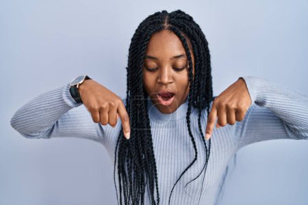 Foto de African american woman standing over blue background pointing down with fingers showing advertisement, surprised face and open mouth - Imagen libre de derechos