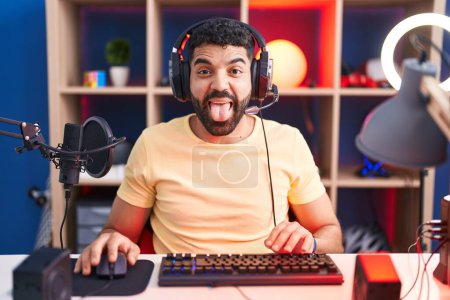 Téléchargez les photos : Hispanic man with beard playing video games with headphones sticking tongue out happy with funny expression. emotion concept. - en image libre de droit