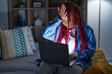Foto de Mujer afroamericana con cabello trenzado usando computadora portátil en la noche sorprendida con la mano en la cabeza por error, recuerde el error. olvidado, mal concepto de memoria. - Imagen libre de derechos