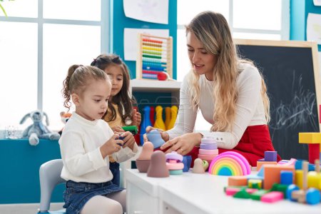 Téléchargez les photos : Teacher with girls playing with geometry blocks sitting on table at kindergarten - en image libre de droit