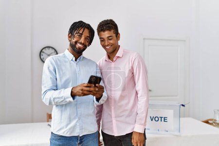 Foto de Dos hombres sonriendo confiados usando smartphone en colegio electoral - Imagen libre de derechos