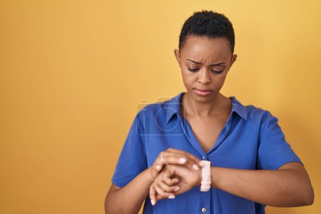Photo for African american woman standing over yellow background checking the time on wrist watch, relaxed and confident - Royalty Free Image