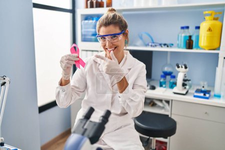 Foto de Young woman working at scientist laboratory holding pink ribbon smiling happy pointing with hand and finger - Imagen libre de derechos