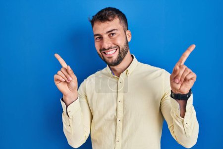 Téléchargez les photos : Bel homme hispanique debout sur fond bleu souriant confiant pointant avec les doigts vers différentes directions. espace de copie pour la publicité - en image libre de droit