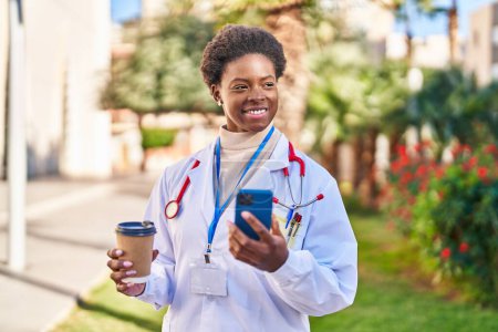 Foto de Mujer afroamericana vistiendo uniforme médico usando teléfono inteligente bebiendo café en el parque - Imagen libre de derechos