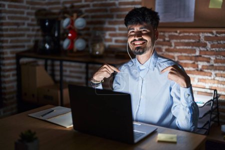 Téléchargez les photos : Jeune homme hispanique avec la barbe travaillant au bureau la nuit regardant confiant avec le sourire sur le visage, se montrant du doigt fier et heureux. - en image libre de droit
