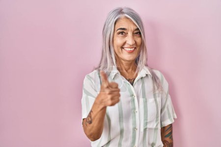 Foto de Middle age woman with grey hair standing over pink background smiling happy and positive, thumb up doing excellent and approval sign - Imagen libre de derechos