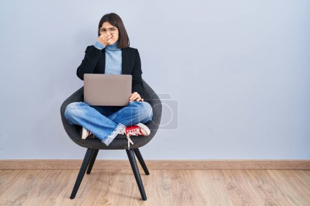 Foto de Young hispanic woman sitting on chair using computer laptop smelling something stinky and disgusting, intolerable smell, holding breath with fingers on nose. bad smell - Imagen libre de derechos