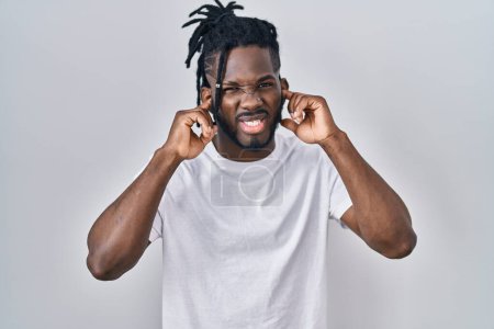 Foto de Hombre africano con rastas con camiseta casual sobre fondo blanco cubriendo las orejas con los dedos con expresión molesta por el ruido de la música alta. concepto de sordo. - Imagen libre de derechos