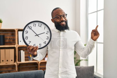 Photo for African american man holding big clock at the office smiling happy pointing with hand and finger to the side - Royalty Free Image