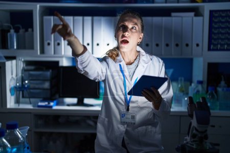 Foto de Beautiful blonde woman working at scientist laboratory late at night pointing with finger surprised ahead, open mouth amazed expression, something on the front - Imagen libre de derechos