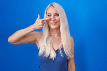Téléchargez les photos : Femme blanche debout sur fond bleu pointant avec le doigt de la main vers le visage et le nez, souriant gai. concept de beauté - en image libre de droit