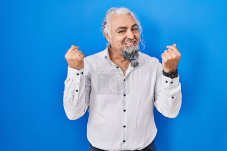 Foto de Middle age man with grey hair standing over blue background doing money gesture with hands, asking for salary payment, millionaire business - Imagen libre de derechos