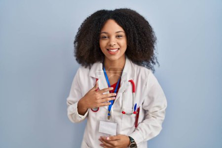 Téléchargez les photos : Jeune femme afro-américaine portant l'uniforme de médecin et stéthoscope souriant et riant fort à haute voix parce que drôle blague folle avec les mains sur le corps. - en image libre de droit