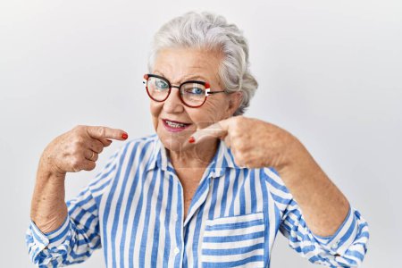 Foto de Mujer mayor con el pelo gris de pie sobre fondo blanco sonriendo alegre mostrando y señalando con los dedos los dientes y la boca. concepto de salud dental. - Imagen libre de derechos