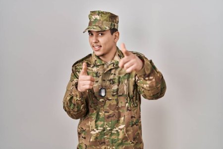 Foto de Hombre árabe joven con camuflaje uniforme del ejército señalando los dedos a la cámara con la cara feliz y divertida. buena energía y vibraciones. - Imagen libre de derechos