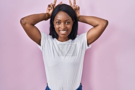 Foto de African young woman wearing casual white t shirt posing funny and crazy with fingers on head as bunny ears, smiling cheerful - Imagen libre de derechos