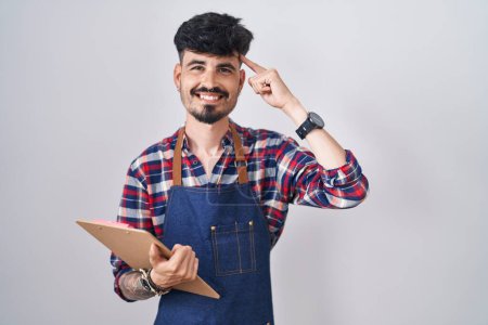 Foto de Young hispanic man with beard wearing waiter apron holding clipboard smiling pointing to head with one finger, great idea or thought, good memory - Imagen libre de derechos