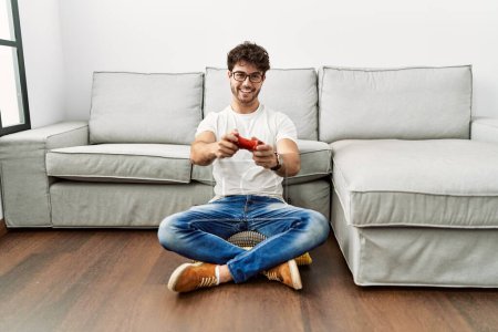 Foto de Joven hombre hispano sonriendo confiado jugando videojuego en casa - Imagen libre de derechos