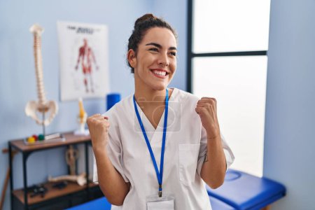 Photo for Young hispanic woman working at rehabilitation clinic very happy and excited doing winner gesture with arms raised, smiling and screaming for success. celebration concept. - Royalty Free Image