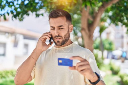 Téléchargez les photos : Young hispanic man talking on smartphone holding credit card at park - en image libre de droit