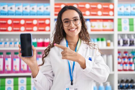 Foto de Mujer hispana joven que trabaja en la farmacia mostrando la pantalla de un teléfono inteligente expresión despistada y confusa. concepto de duda. - Imagen libre de derechos