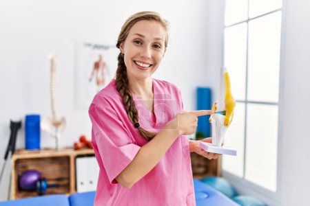 Téléchargez les photos : Jeune femme caucasienne portant l'uniforme de physiothérapeute tenant le modèle anatomique du genou à la clinique de physiothérapie - en image libre de droit