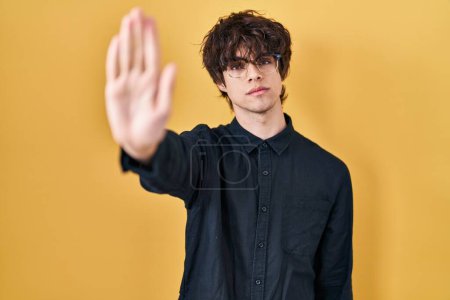Foto de Young man wearing glasses over yellow background doing stop sing with palm of the hand. warning expression with negative and serious gesture on the face. - Imagen libre de derechos