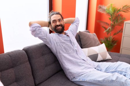Foto de Joven hombre hispano relajado con las manos en la cabeza sentado en el sofá en casa - Imagen libre de derechos