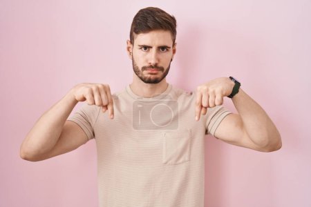 Téléchargez les photos : Hispanic man with beard standing over pink background pointing down looking sad and upset, indicating direction with fingers, unhappy and depressed. - en image libre de droit