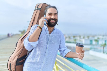 Téléchargez les photos : Young hispanic man musician talking on smartphone drinking coffee at seaside - en image libre de droit