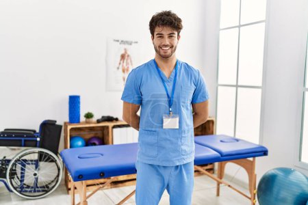 Téléchargez les photos : Jeune homme hispanique portant un uniforme de physiothérapeute debout à la clinique - en image libre de droit