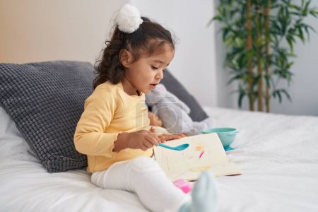 Foto de Adorable niño hispano sentado en la cama leyendo libro en el dormitorio - Imagen libre de derechos