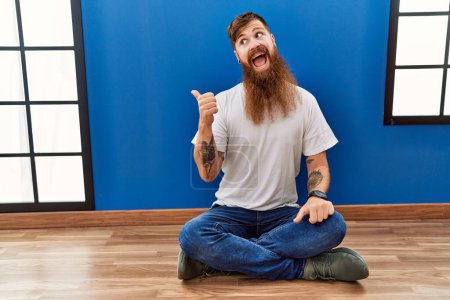 Foto de Redhead man with long beard sitting on the floor at empty room smiling with happy face looking and pointing to the side with thumb up. - Imagen libre de derechos