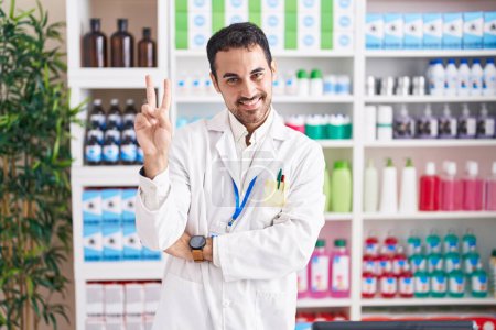 Téléchargez les photos : Bel homme hispanique travaillant à la pharmacie pharmacie souriant avec un sourire heureux clin d'œil à la caméra faisant signe de victoire avec les doigts. numéro deux. - en image libre de droit