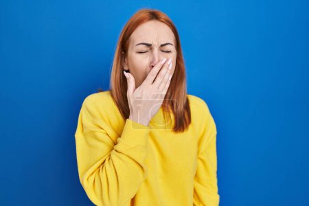 Foto de Young woman standing over blue background bored yawning tired covering mouth with hand. restless and sleepiness. - Imagen libre de derechos