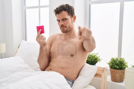 Photo for Young hispanic man holding condom in the bed pointing with finger to the camera and to you, confident gesture looking serious - Royalty Free Image