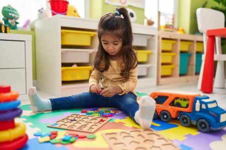 Foto de Adorable chica hispana jugando con las matemáticas juego de puzzle sentado en el suelo en el jardín de infantes - Imagen libre de derechos