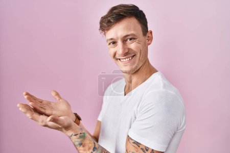 Téléchargez les photos : Homme caucasien debout sur fond rose pointant de côté avec les mains ouvertes paumes montrant l'espace de copie, présentant la publicité souriant heureux excité - en image libre de droit
