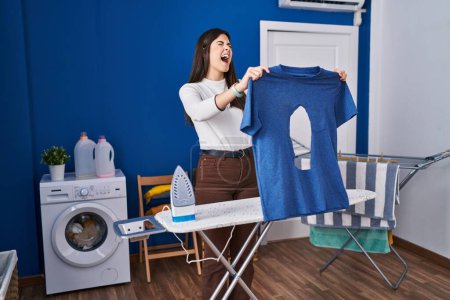 Téléchargez les photos : Young brunette woman ironing holding burned iron shirt at laundry room angry and mad screaming frustrated and furious, shouting with anger looking up. - en image libre de droit